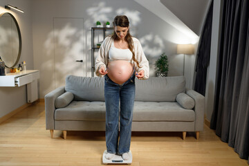Fototapeta premium Expecting mother stands on scale in living room, checking weight gain during pregnancy, feeling clothes tightness