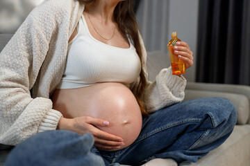 Pregnant woman sits on sofa holding bottle of oil for belly care during pregnancy
