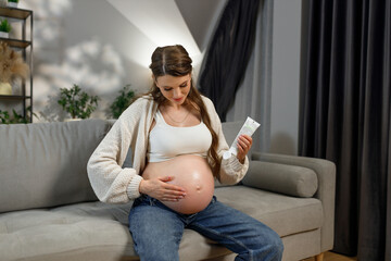 Pregnant woman sitting on sofa applying cream to her belly for skin care during pregnancy