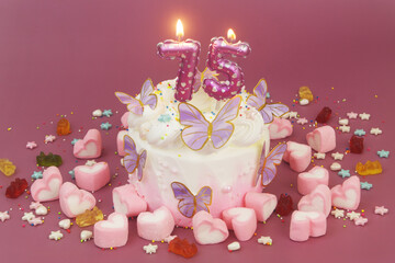Birthday cake with butterflies, air balloon shaped candles number 75, jelly and marshmallow candies on purple background.