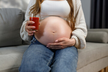 Third trimester pregnant woman sits on sofa holding glass of tomato juice, cradling her belly with her hands