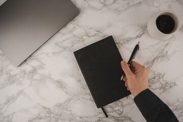 A person writes in a black notebook with a pen on a white marble desk. A laptop and a cup of coffee are also visible, suggesting a casual workspace