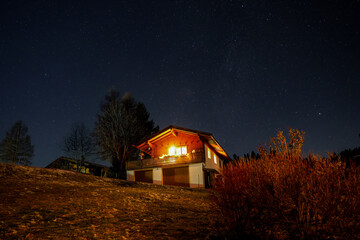 Illuminated chalet-style house surrounded by trees and bushes at night under a clear starry sky. The building is a traditional wooden chalet with a gabled roof and an illuminated balcony