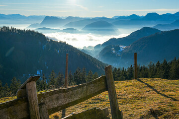 An old, weathered wooden fence in a meadow overlooking a breathtaking mountain landscape in the Chiemgau Alps with snow-covered slopes and fog in the valleys and the sun high in the sky.