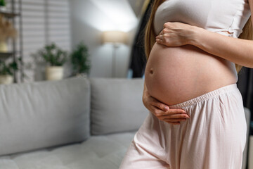 Pregnant woman in light pajamas gently cradles her belly while standing indoors near a sofa and plants