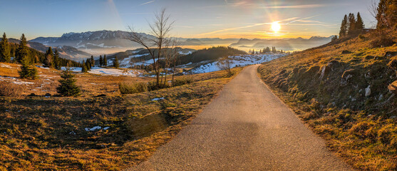 Picturesque winter landscape in the Chiemgau Alps, where a mountain road winds through a hilly landscape with snow-covered slopes. A brilliant sunset bathes the mountains in warm, golden light.