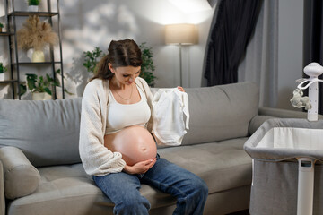Expecting mother sits on sofa holding tiny white baby onesie, preparing for newborn arrival with crib nearby