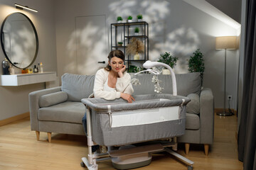 Woman sits by bedside bassinet with mobile, contemplating motherhood in a modern living room setting