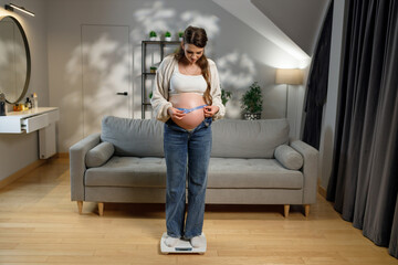 Expecting mother stands on scale measuring her pregnant belly with tape measure in living room