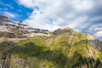 Fototapeta premium Sharp green ridges and rugged mountain peaks rise beneath a partly cloudy sky in the Pyrenees, with sunlight casting dynamic shadows across the rocky slopes.