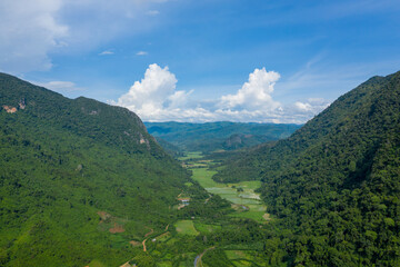 Fototapeta premium Wide aerial view of a vibrant green valley surrounded by steep forested hills in northern Laos. The landscape features patchwork rice fields, dense jungle, and a bright blue sky with dramatic cloud