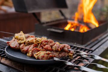 Naklejka premium Raw skewers of meat laid out on dark plate, open fire in a grill in the background, grilling, preparing food on backyard