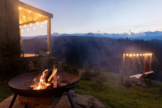 Campfire burning in round metal pit, house with mountains view on background, smoke rising from the fire, blending with the evening