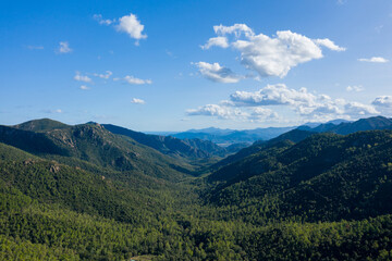 Fototapeta premium Aerial view of a vast green valley surrounded by rugged mountains and dense woodland in Sardinia, Italy. Puffy clouds drift across a clear blue sky, casting gentle shadows over the landscape.