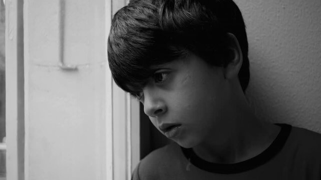 Child looking out window with lowered gaze in quiet moment suggesting loneliness sadness introspection early emotional struggle childhood melancholy and reflective inner tension in black and white