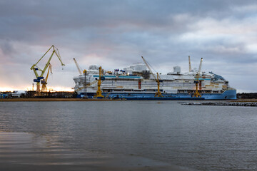 Photo of a large ship being constructed at a shipyard near the sea, captured from a distant vantage point. Shows cranes, scaffolding, massive vessel structure, and industrial maritime environment.