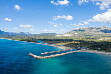 Obraz premium Aerial image of stone breakwaters extending into clear turquoise Mediterranean waters at the entrance of Orosei marina, with a sweeping sandy coastline and rugged mountains under a bright sky.