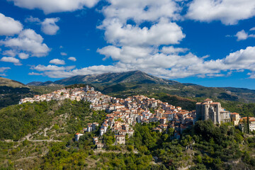Obraz premium Wide aerial view of Rivello, Italy, featuring clustered terracotta rooftops atop a green hillside, backed by dramatic mountains under a sky filled with scattered clouds. Crisp daylight enhances the