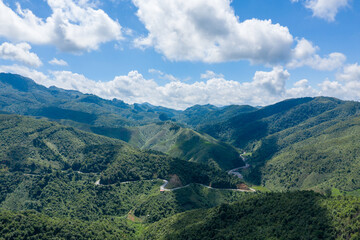 Fototapeta premium Sweeping aerial view of a winding mountain road cutting through lush green hills and valleys under a bright blue sky with scattered clouds in northern Laos. The landscape features layered ridges