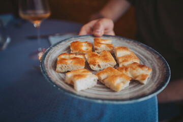 A person holds a plate of freshly baked bread pieces in a restaurant. The setting includes a glass of white wine nearby on the table. Soft light creates a cozy atmosphere