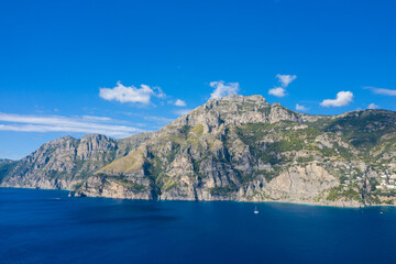 Obraz premium Sheer rocky mountains rise from vibrant blue waters under a clear sky, with scattered white clouds and sunlight highlighting the rugged coastline. The scene captures the dramatic landscape and natural