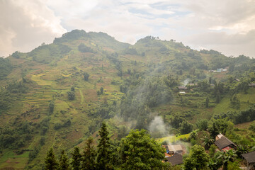 Gentle haze drifts over green hillsides dotted with terraced rice fields and rural houses near Bac Ha, Vietnam. Soft afternoon light and drifting smoke create a tranquil, layered landscape.