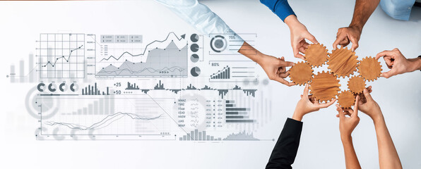 A diverse group of professionals collaborates over a table, joining hands around gears while surrounded by business graphs and data visuals, representing teamwork and strategic planning. Scalp