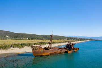 Corroded remains of the Dimitrios shipwreck rest in shallow turquoise water near a sandy coastline outside Gythio, with green hills and a bright blue sky creating a striking contrast. The midday sun