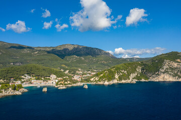 Obraz premium Wide view of the Parga coastline in Epirus, Greece, featuring deep blue water, rocky cliffs, and a coastal village backed by densely forested hills. Bright daylight and scattered clouds enhance the