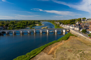 Fototapeta premium High-angle perspective of the historic stone bridge spanning the Loire river in La Charite sur Loire, with traditional rooftops lining the riverbank and dense green forest beyond. The scene captures