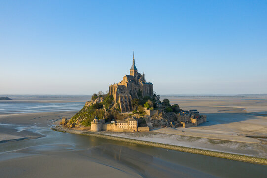 Early morning light bathes Mont-Saint-Michel and its medieval stone architecture, with expansive tidal sands and tranquil water channels stretching into the distance beneath a clear blue sky.