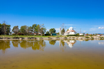 Obraz premium A white-domed church and a row of green trees are reflected in the calm water of Tourlida lagoon in western Greece. The scene features clear blue sky, tranquil wetland, and natural shoreline textures.