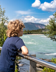 A boy with blond curly hair stands by a turquoise river in Norway, enjoying the sunny day during his summer vacation. He gazes at the water and the distant mountains