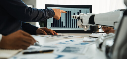 A business team collaborates in a modern office setting, analyzing data presented on a screen, with a focus on trends and strategic planning aided by robotic technology.Noogenesis