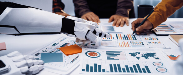 A robotic hand assists a business team analyzing data and developing strategies through charts and graphs, showcasing the blend of technology and teamwork in modern offices.Noogenesis