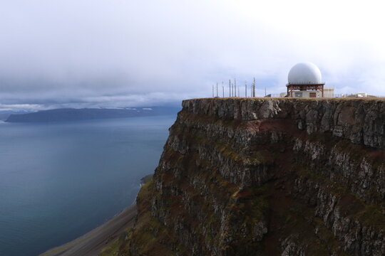 Radar dome and communication towers perched on a dramatic coastal cliff overlooking the ocean, combining advanced technology with rugged natural scenery under an overcast sky