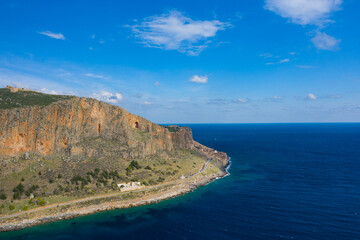 Obraz premium Steep orange cliffs of Monemvasia overlook a winding coastal road and vivid turquoise sea beneath a wide blue sky. The dramatic landscape is bathed in sunlight, highlighting rocky textures and natural