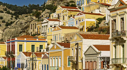 view of Symi, Greece, showcasing a cluster of colorful buildings stacked along a rocky slope. The architecture features pastel facades with tiled roofs and shuttered windows