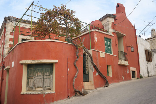 Vibrant red traditional house on Mandilara Street in Archanes, Crete. Features a thick vine climbing the facade, green wooden shutters and classic Mediterranean charm.