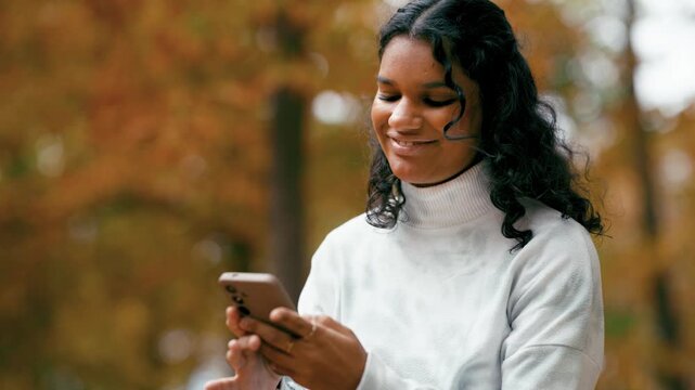 Cheerful young woman with long curly hair using her smartphone and smiling. Beautiful natural background with golden autumn leaves and trees
