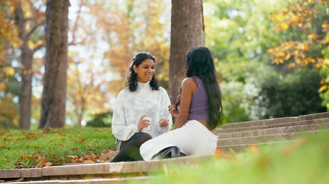Two cheerful young indian women sitting on stairs in an autumn park. They are laughing and talking while enjoying a sunny day outdoors