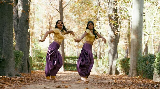 Two joyful indian women wearing traditional costumes dancing a synchronized choreography in an autumn park. Their energetic moves showcase india's rich culture