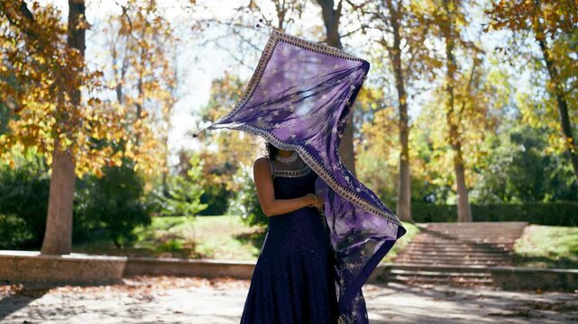 Beautiful young woman performing a traditional indian dance in a sunny park. Graceful dancer smiling and spinning with a flowing purple sari