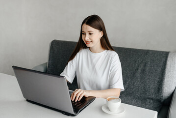 young beautiful caucasian brunette woman freelancer works in cafe with notebook, casual clothes, white T-shirt, cup of coffee, grey couch and notebook