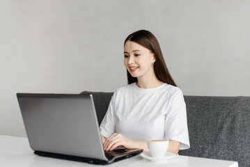 young beautiful caucasian brunette woman freelancer works in cafe with notebook, casual clothes, white T-shirt, cup of coffee, grey couch and notebook