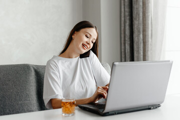 young beautiful caucasian brunette woman freelancer works in cafe with notebook, casual clothes, white T-shirt, apple juice in glass, grey couch and notebook, talking by phone, multy-tasking