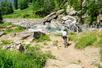 Mountain biker riding on a dirt trail near a clear alpine stream and small waterfall, surrounded by green forest and rocks on a sunny summer day in the mountains.