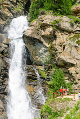 Tourists admiring the powerful Lillaz waterfalls near Cogne, Italy, with cascading alpine water, rocky cliffs and lush greenery on a bright summer day in the mountains.