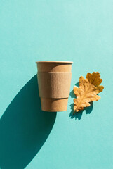 Eco coffee cup and autumn leaf against a teal background. Kraft coffee cup and dry autumn oak leaf flat lay on turquoise background.