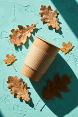 Eco coffee cup and autumn leaf against a teal background. Disposable coffee cup and autumn dry leaves on bright textured teal background.
